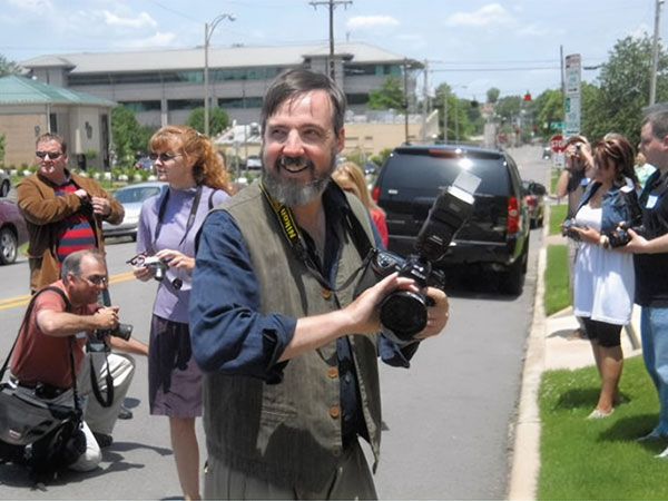 Jack standing on a street smiling looking to the side while holding a camera as other photographers take pictures nearby